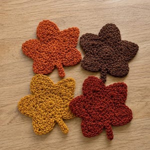 May include: Four crochet leaf-shaped coasters in autumn colors. The set includes one orange, one dark brown, one mustard yellow, and one deep red leaf, each featuring a textured, intricate stitch pattern and a small stem at the base.
