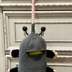 Puede incluir: Una bolsa de ganchillo con forma de criatura gris, con un lazo rosa para colgar, cuatro extremidades negras con borde rosa y una abertura blanca en la parte inferior. El cuerpo tiene una ranura horizontal que muestra una bolsa amarilla.