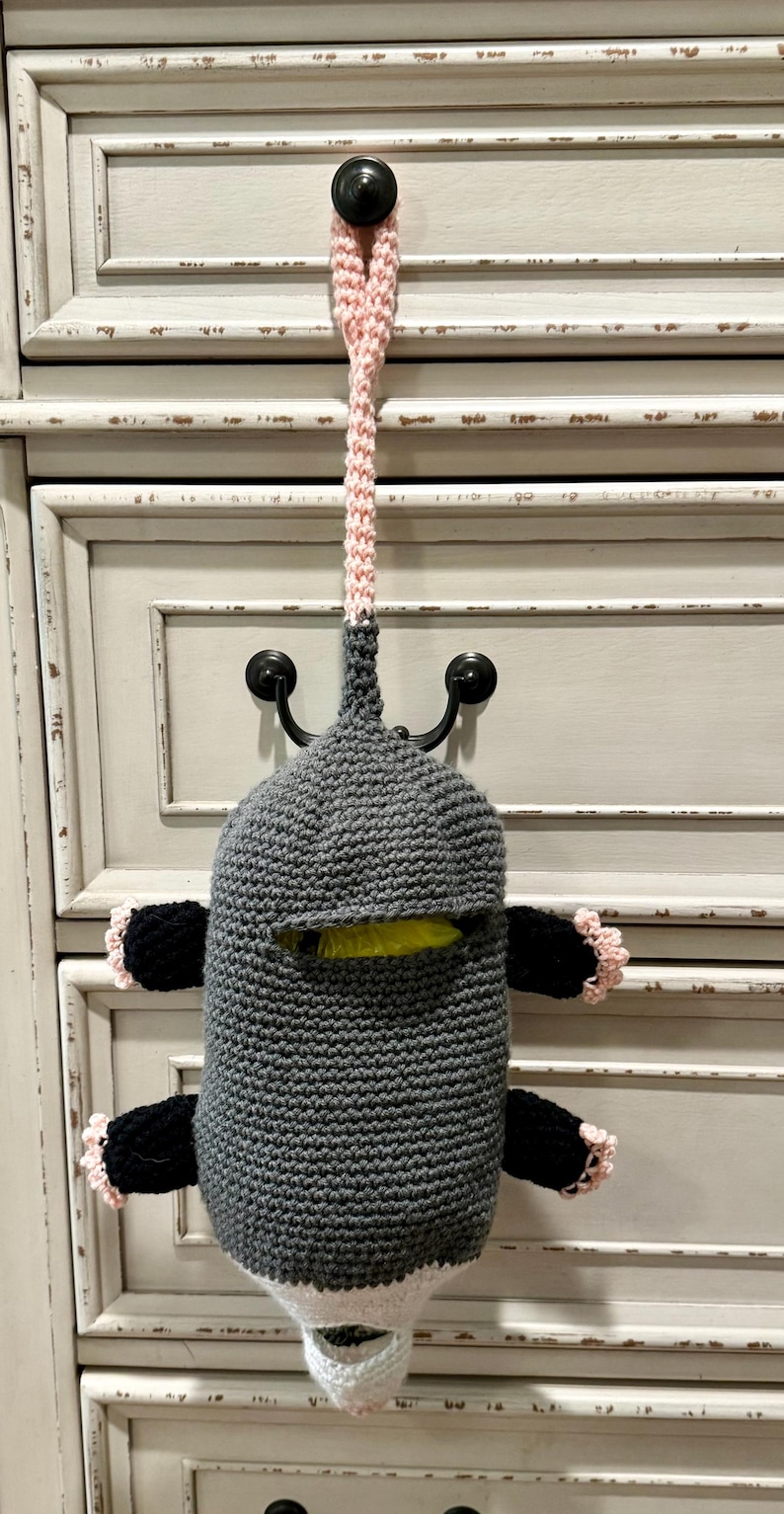 Puede incluir: Una bolsa de ganchillo con forma de criatura gris, con un lazo rosa para colgar, cuatro extremidades negras con borde rosa y una abertura blanca en la parte inferior. El cuerpo tiene una ranura horizontal que muestra una bolsa amarilla.