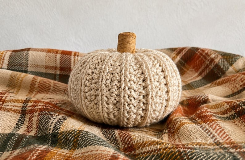May include: A round, cream-colored crocheted pumpkin with a textured, ribbed surface and a natural brown cork stem. The pumpkin features a dense, hand-stitched yarn construction with vertical ridges that mimic the appearance of a real gourd.