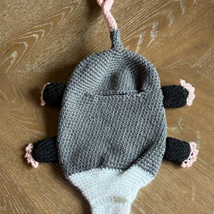 Puede incluir: Una toalla o bolsa colgante de ganchillo con forma de zarig&uuml;eya que presenta un cuerpo gris, una cara blanca con nariz rosa, cuatro extremidades negras con garras de punta rosa y un lazo de cola rosa en la parte superior. Incluye una abertura.