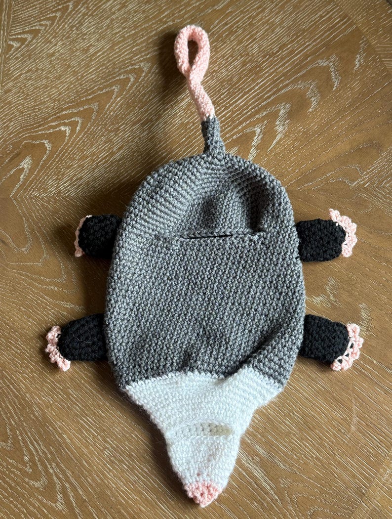 Puede incluir: Una toalla o bolsa colgante de ganchillo con forma de zarig&uuml;eya que presenta un cuerpo gris, una cara blanca con nariz rosa, cuatro extremidades negras con garras de punta rosa y un lazo de cola rosa en la parte superior. Incluye una abertura.