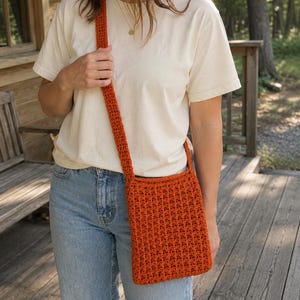 Puede incluir: Un bolso bandolera de ganchillo en color naranja quemado con un patrón de puntos texturizado y repetitivo, y una correa larga a juego. El bolso tiene forma rectangular, está hecho de hilo y presenta un tejido denso y consistente.
