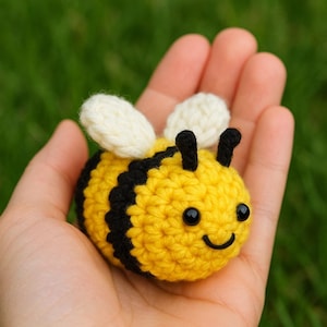 May include: A crocheted bee toy, featuring yellow and black stripes, white wings, and a smiling face. The bee is held in an open hand. The background is a blurred green.