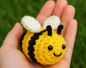 May include: A handmade crochet bee with yellow and black stripes, white wings, and black eyes. The bee is smiling and held in an open hand. The background is blurred green.