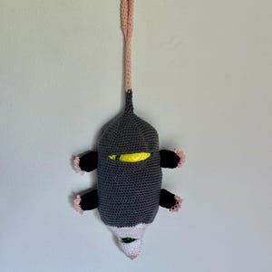Puede incluir: Un dispensador de bolsas de ganchillo con forma de zarig&uuml;eya, cuerpo gris oscuro, extremidades negras con borde rosa claro, cara blanca con nariz rosa y una larga cola rosa claro en forma de bucle para colgar. Se ve una bolsa amarilla dentro.