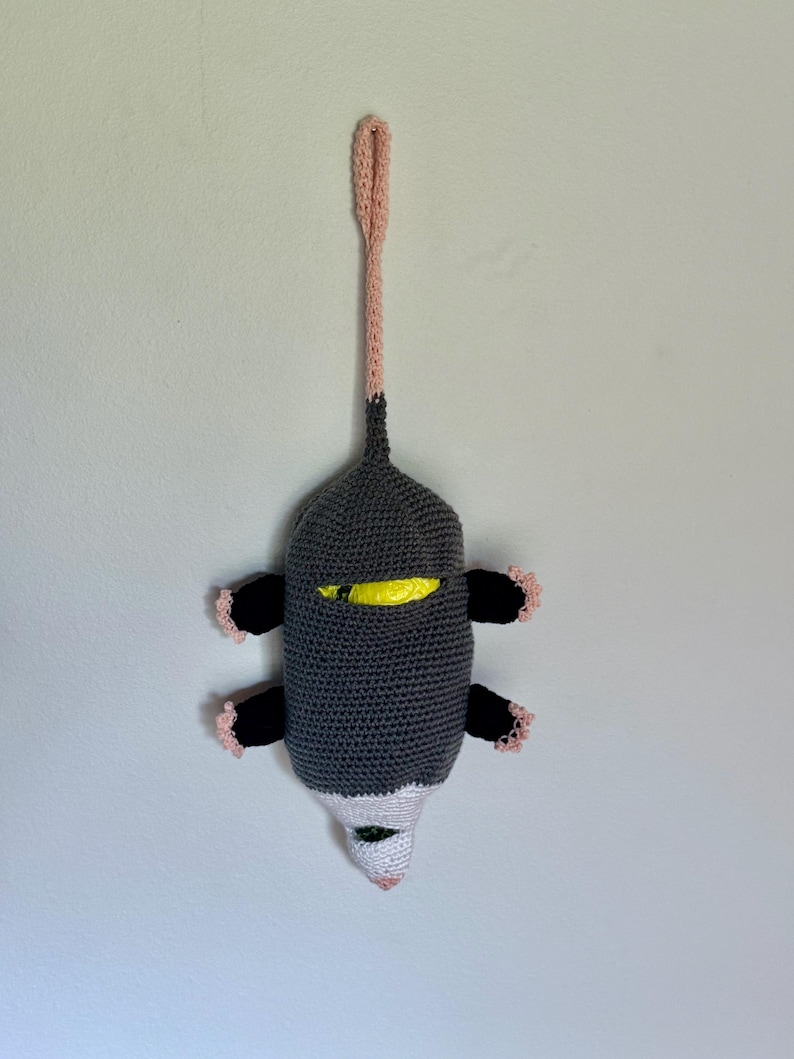 Puede incluir: Un dispensador de bolsas de ganchillo con forma de zarig&uuml;eya, cuerpo gris oscuro, extremidades negras con borde rosa claro, cara blanca con nariz rosa y una larga cola rosa claro en forma de bucle para colgar. Se ve una bolsa amarilla dentro.