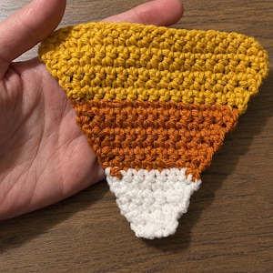 May include: A triangular crochet piece shaped like a candy corn, featuring horizontal bands of yellow at the top, orange in the middle, and white at the bottom. The texture is created with dense, uniform stitches, forming a flat, pointed shape.
