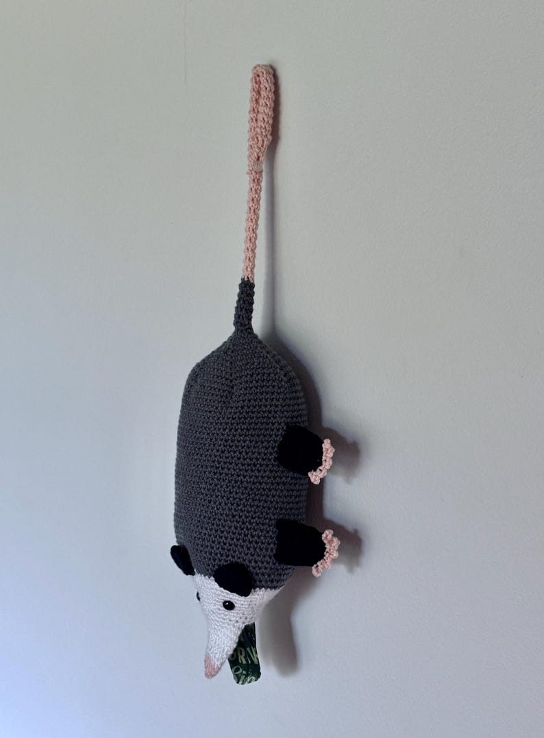 Puede incluir: Un juguete de peluche de zarig&uuml;eya tejido a ganchillo con cuerpo gris oscuro, extremidades negras y cara blanca con peque&ntilde;os ojos negros. La zarig&uuml;eya tiene una cola larga tejida en rosa claro hacia arriba y un trozo de tela verde en la boca.