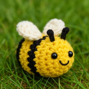 May include: A handmade crochet bee with yellow and black stripes, cream-colored wings, and black eyes. The bee is smiling and sits in a bed of green grass. This item is a cute, handcrafted toy.