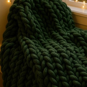 May include: A chunky knit blanket in a deep forest green color. The blanket features a thick, hand-knitted texture with large, interlocking loops of soft yarn, creating a dense and heavy appearance throughout the entire piece of textile.