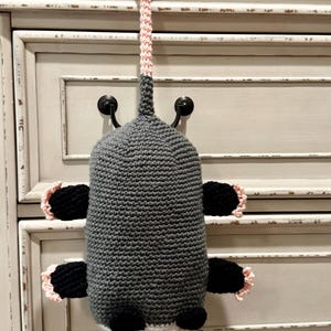Puede incluir: Un juguete de zarig&uuml;eya tejido a ganchillo con cuerpo gris, cara blanca con ojos negros y extremidades negras con detalles en rosa claro. El juguete incluye una cola gris larga y texturizada que termina en un lazo rosa para colgar.