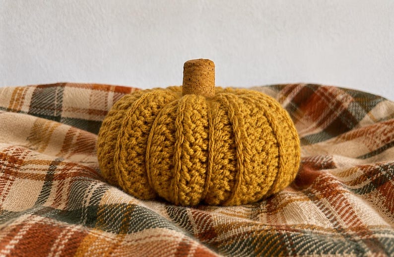 May include: A mustard yellow crocheted pumpkin featuring a textured ribbed pattern and a small, cylindrical cork stem attached to the top center. The pumpkin has a rounded, segmented shape created by vertical stitches that define its form.