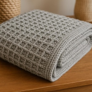 May include: A folded, gray knitted blanket with a waffle-like pattern. The blanket is made of a soft, textured material and has a thick border. The blanket is folded neatly and appears to be new.