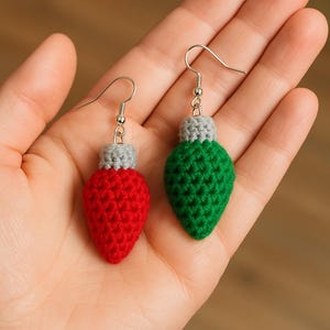 Puede incluir: Un par de pendientes de ganchillo hechos a mano con forma de bombillas navideñas. Un pendiente es rojo, el otro es verde, ambos con la parte superior gris y ganchos plateados. Los pendientes se sostienen en una mano.