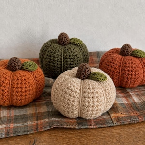 May include: Four handmade crochet pumpkins in varying colours including orange, olive green, and cream. Each pumpkin features a textured, ribbed surface, a small brown crocheted stem at the top, and a single small green crocheted leaf attached to the side.
