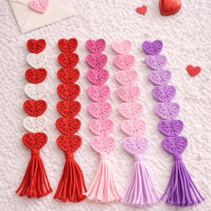 以下が含まれることがあります： 赤、白、ピンク、紫の、かぎ針編みのハートガーランド。それぞれに同じ色のタッセルが付いています。小さなピンクのハートが白い封筒の上にあり、赤いハート型のキャンディーもあります。白いテクスチャのある表面に配置されています。