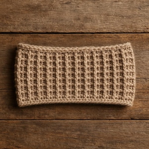 Może przedstawiać: Beżowa dzianinowa opaska na głowę o strukturze przypominającej gofry. Opaska jest prostokątna i wydaje się być wykonana z miękkiej, ciepłej przędzy. Tło stanowi drewniana powierzchnia.