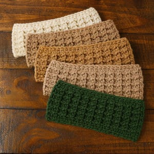May include: Five handmade crochet headbands in varying colors: cream, brown, tan, light brown, and dark green. The headbands are arranged in a diagonal row, showcasing their textured stitch pattern. The headbands are made of soft yarn.