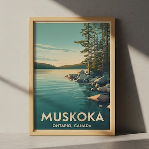 May include: A framed art print showing a tranquil lake scene with the text "MUSKOKA, ONTARIO, CANADA." The artwork depicts a calm lake with ripples, rocky shoreline, and evergreen trees under a blue sky. The frame is a light wood colour.