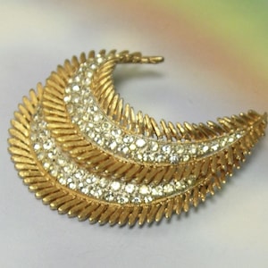 May include: A gold-toned brooch in a crescent shape, featuring three rows of textured, feather-like details. The brooch is embellished with clear, sparkling rhinestones, adding a touch of glamour to the design. The overall aesthetic is vintage and elegant.