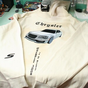 Puede incluir: Sudadera con capucha color crema con la palabra "Chrysler" en escritura negra sobre un gráfico de coche. El coche es gris y blanco con detalles en azul. La manga tiene un logotipo "S" negro.