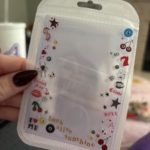 Puede incluir: Portatarjetas de plástico transparente decorado con varios símbolos, incluyendo cerezas, estrellas y palos de cartas. Las palabras "Look Alive Sunshine" están impresas en el soporte. El soporte está en un paquete de plástico transparente.