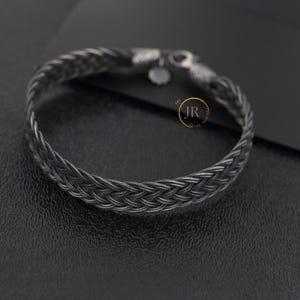 May include: A dark gray braided bracelet featuring a woven metal chain design with a lobster claw clasp and a small circular charm. The bracelet displays a circular logo with the letters JR in the center, positioned near the clasp mechanism.