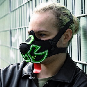 May include: Black and green dog-themed mask with a neon green outline and a red tongue. The mask is worn with a black strap. The person is wearing a black leather jacket.