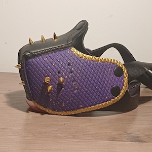 May include: A black and purple leather mask with gold-coloured accents and spikes. The mask features a quilted purple centre panel, gold trim, and gold-coloured spikes along the top edge. Black straps and fasteners complete the design.