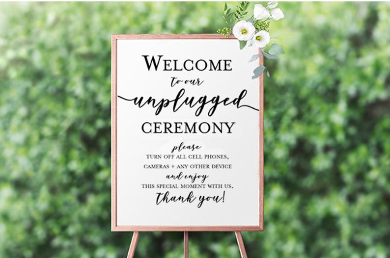May include: A white sign with a wooden frame on an easel, announcing an unplugged ceremony. The sign reads "Welcome to our unplugged ceremony, please turn off all cell phones, cameras and any other device and enjoy this special moment with us. Thank you!"