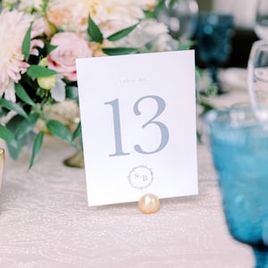 以下が含まれることがあります： 白いカードにグレーの文字で「table no. 13」と書かれています。カードは小さな金の真珠で支えられています。カードには、中央に「S+B」のイニシャルが入った小さな花輪があります。