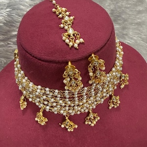 May include: A gold-toned necklace, earrings, and headpiece set. The jewelry features small white pearls and clear, round gemstones. The necklace and headpiece have a delicate, layered design. The set is displayed on a maroon surface.