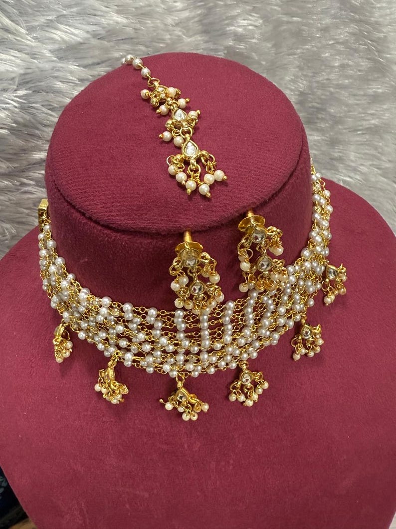 May include: A gold-toned necklace, earrings, and headpiece set. The jewelry features small white pearls and clear, round gemstones. The necklace and headpiece have a delicate, layered design. The set is displayed on a maroon surface.