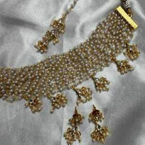 May include: An elegant jewelry set featuring a multi-strand pearl necklace, matching earrings, and a headpiece. The set has gold-toned accents and decorative pendants. The pearls are white, and the overall design is intricate and ornate.