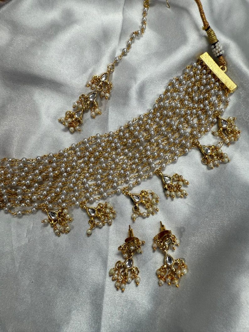 May include: An elegant jewelry set featuring a multi-strand pearl necklace, matching earrings, and a headpiece. The set has gold-toned accents and decorative pendants. The pearls are white, and the overall design is intricate and ornate.
