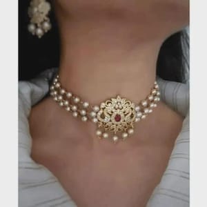 May include: A gold choker necklace with three strands of pearls. The central pendant has an ornate design with a red gemstone and clear accents. Matching earrings are visible. The jewellery is likely for a special occasion.