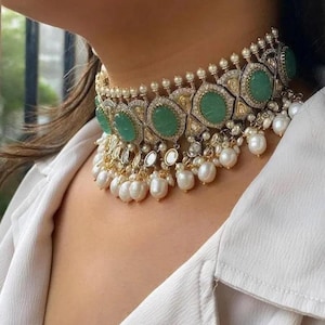 Könnte beinhalten: Ein goldfarbenes Choker-Halsband mit ovalen grünen Edelsteinen und kleinen, runden Perlen. Die Halskette hat ein detailliertes Design mit klaren, funkelnden Akzenten und hängenden Perltropfen, was einen eleganten und verzierten Look erzeugt.