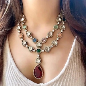 May include: A two-strand necklace with a variety of colourful gemstones. The necklace features a teardrop-shaped, deep red gemstone pendant. The gemstones are set in a gold-coloured metal and are surrounded by smaller, clear stones.