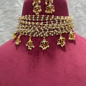 May include: A gold-toned necklace and earring set. The necklace is a choker style, featuring multiple strands of small, white pearls. Gold pendants with pearl accents hang from the necklace. The earrings are a matching design, with gold and pearl details.