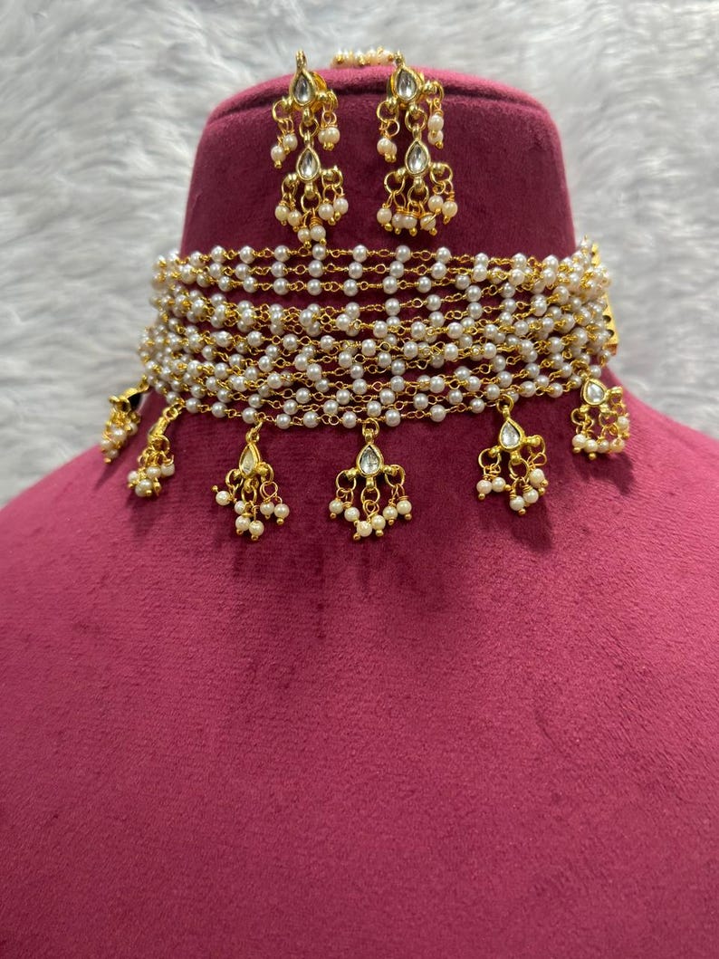 May include: A gold-toned necklace and earring set. The necklace is a choker style, featuring multiple strands of small, white pearls. Gold pendants with pearl accents hang from the necklace. The earrings are a matching design, with gold and pearl details.