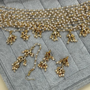 May include: A gold-toned jewelry set featuring a multi-strand pearl necklace with dangling pendants, a matching headpiece, and earrings. The set includes clear gemstone accents and small pearl clusters, displayed on a gray surface.