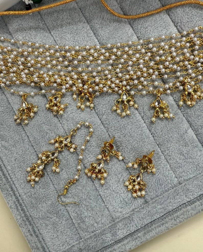 May include: A gold-toned jewelry set featuring a multi-strand pearl necklace with dangling pendants, a matching headpiece, and earrings. The set includes clear gemstone accents and small pearl clusters, displayed on a gray surface.