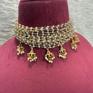 May include: A gold-toned choker necklace with multiple strands of small, white pearls. The necklace has dangling gold pendants with pearl and gemstone accents. The necklace is displayed on a maroon bust.