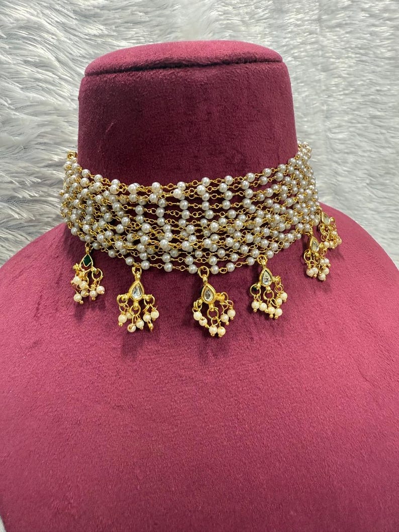 May include: A gold-toned choker necklace with multiple strands of small, white pearls. The necklace has dangling gold pendants with pearl and gemstone accents. The necklace is displayed on a maroon bust.