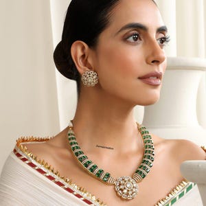 May include: A gold necklace and matching earrings set, featuring green gemstones and a floral design. The necklace has a central floral pendant. The earrings are also floral-shaped. The woman is wearing a white garment with gold trim.