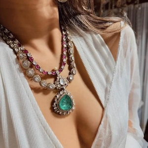 May include: An elaborate necklace and earring set featuring a large emerald pendant. The necklace incorporates clear and ruby-coloured stones, with a matching earring design. The jewellery is set against a white garment, creating a striking contrast.