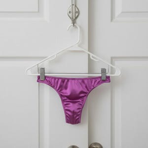Puede incluir: Un tanga brillante de color magenta cuelga de una percha de plástico blanco. La ropa interior está hecha de un material liso similar al satén. La percha está suspendida de un gancho de metal en una puerta blanca.