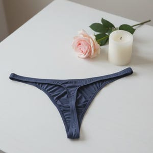 Könnte beinhalten: Dunkelblauer String-Tanga. Eine hellrosa Rose und eine weiße Kerze liegen neben der Unterwäsche. Die Unterwäsche ist aus einem weichen, dünnen Material gefertigt.