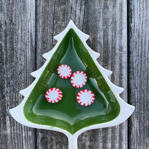 May include: A green ceramic Christmas tree-shaped dish with a white rim, filled with four red and white peppermint candies.